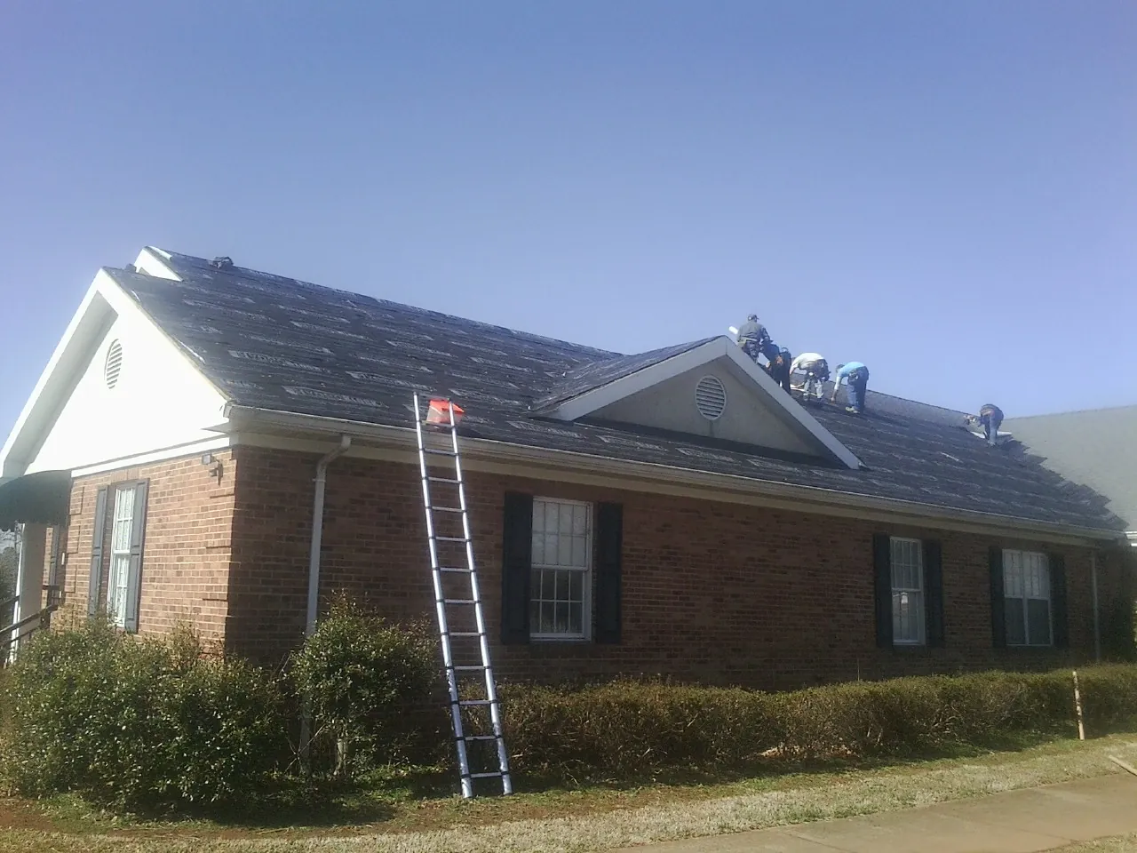 Crew tearing shingles off a brick ranch roof with a ladder set — Emergency Roof Repair work by our crew in Fork